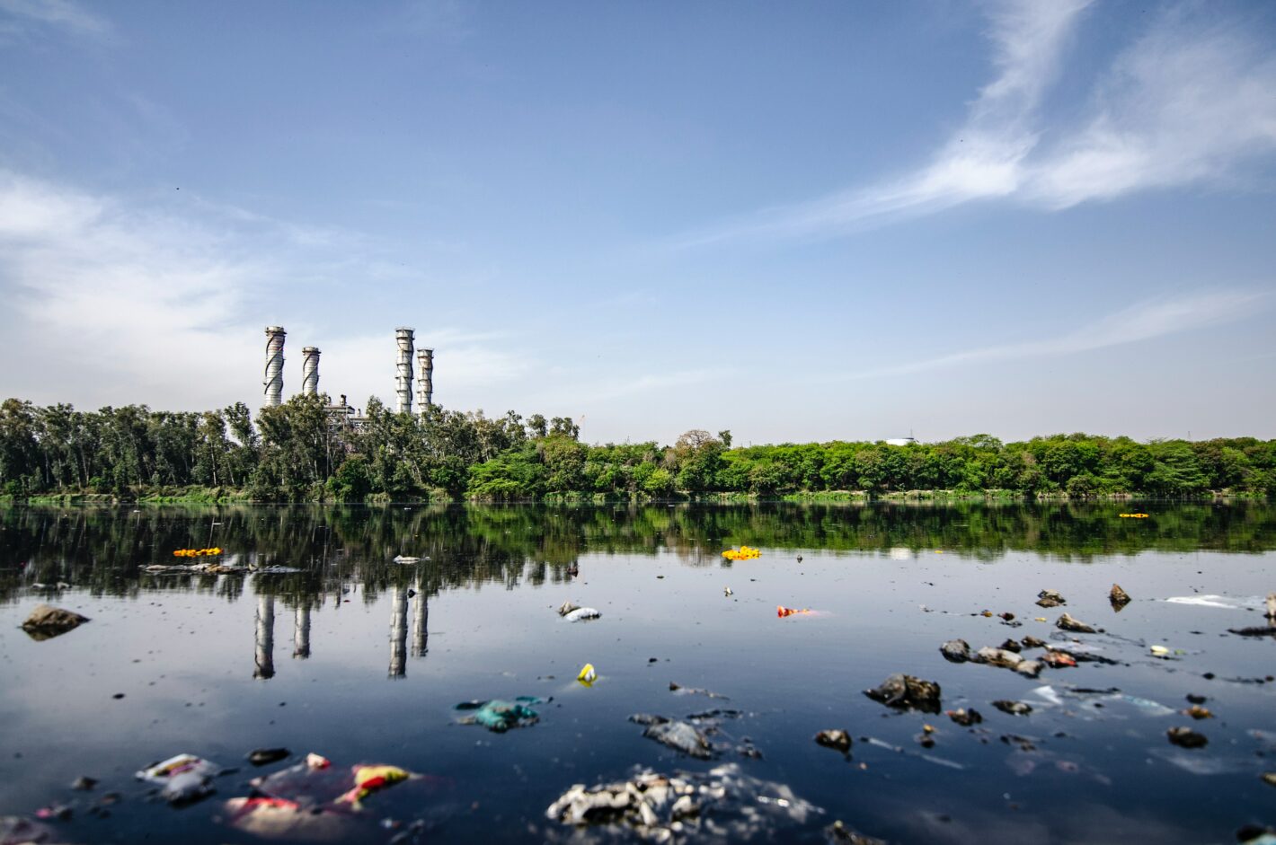 Mit Plastikmüll verschmutztes Gewässer. Im Hintergrund sind Schornsteine einer Fabrik zu sehen.