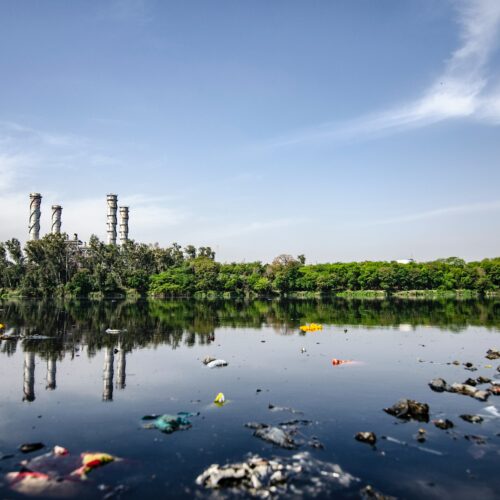 Mit Plastikmüll verschmutztes Gewässer. Im Hintergrund sind Schornsteine einer Fabrik zu sehen.